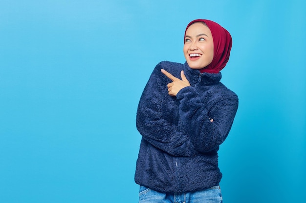 Cheerful young Asian woman pointing fingers aside at copy space isolated over blue background