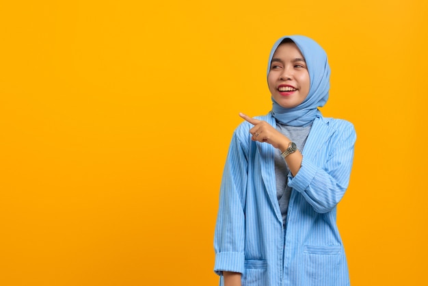 Cheerful young Asian woman pointing fingers aside at copy space isolated over yellow background