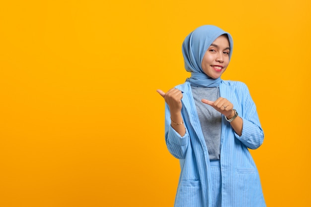 Cheerful young Asian woman pointing two fingers aside at copy space isolated over yellow background