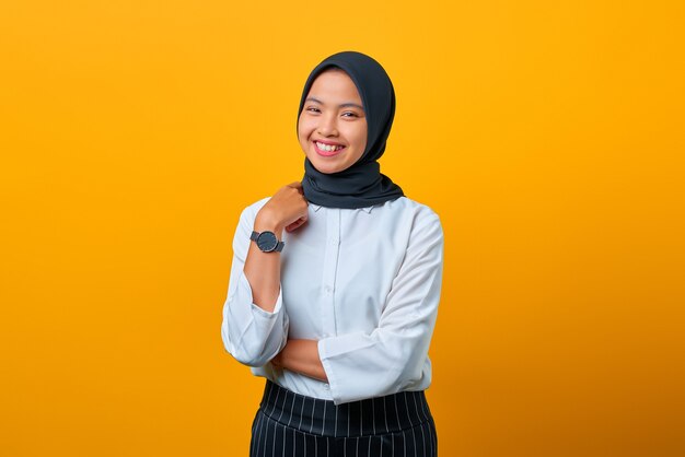 Cheerful young Asian woman raised hand with crossed arm isolated over yellow background