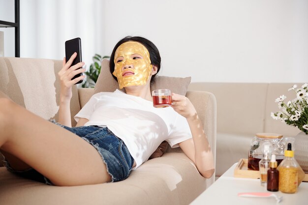 顔にシリコンシートマスクを付けてソファで休んで、ハーブティーを飲み、スマートフォンでソーシャルメディアをチェックする陽気な若いアジアの女性