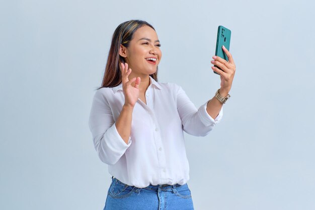 Cheerful young Asian woman saying hi and waving hand while making video call on mobile phone isolated over white background