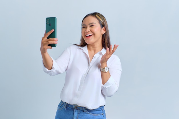 Cheerful young Asian woman saying hi and waving hand while making video call on mobile phone isolated over white background