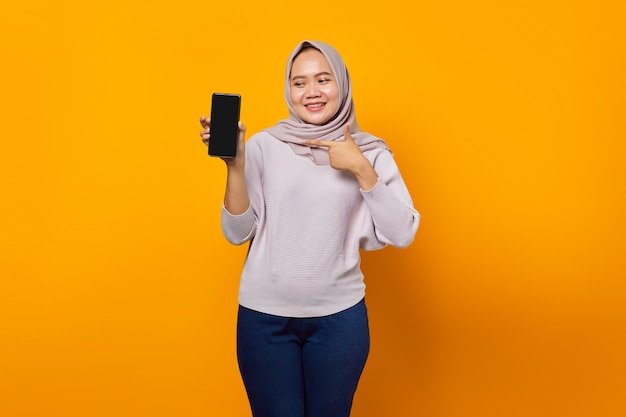 黄色の背景で隔離のスマートフォンの空白の画面を示す陽気な若いアジアの女性