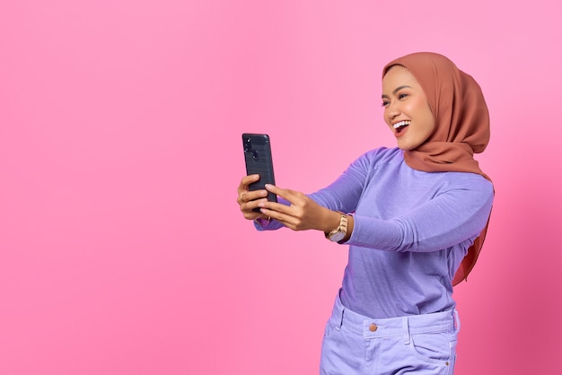 Cheerful young asian woman taking a selfie with a mobile phone
on pink background