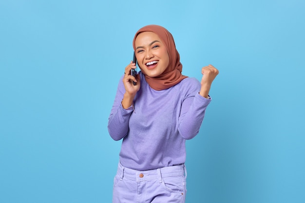 Cheerful young Asian woman talking on mobile phone while celebrating victory on blue background