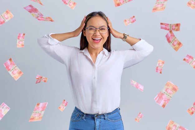 Cheerful young Asian woman touching his head with money banknotes flying in the air isolated over white background