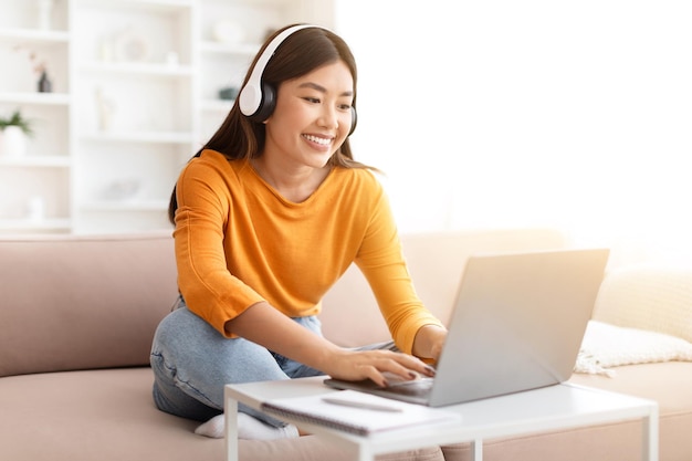 Cheerful young asian woman using laptop and headphones at home