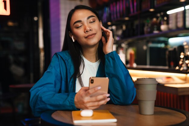 cheerful young asian woman using mobile phone and wireless earphones while sitting in cafe