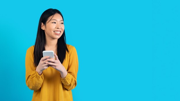 Cheerful young asian woman using phone on blue copy space