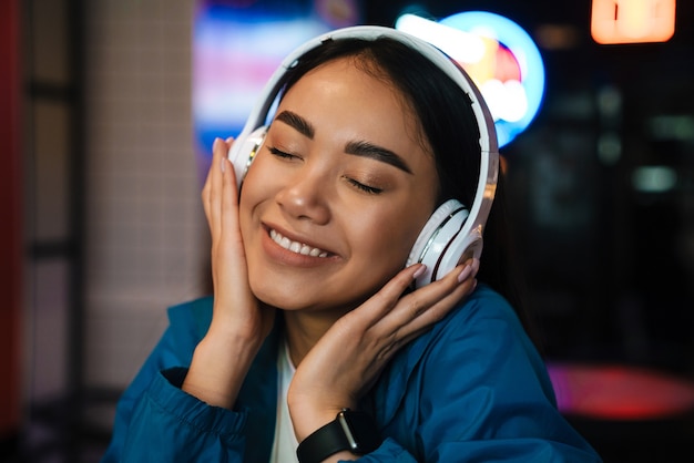 cheerful young asian woman using wireless headphones and smiling while sitting in cafe