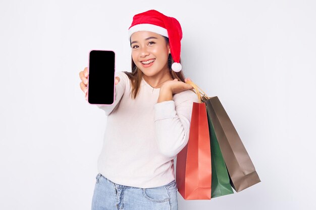 買い物袋を運ぶクリスマス帽子をかぶって、白い背景で隔離の携帯電話で推奨されるクリスマス プロモーションを示す陽気な若いアジア女性