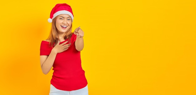 写真 黄色の背景の上のカメラに指を指しているクリスマスの帽子をかぶって陽気な若いアジアの女性