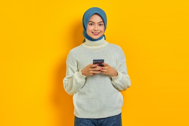 Cheerful young asian woman in white sweater and hijab using on mobile phone and looking at camera isolated over yellow background People islam religious concept