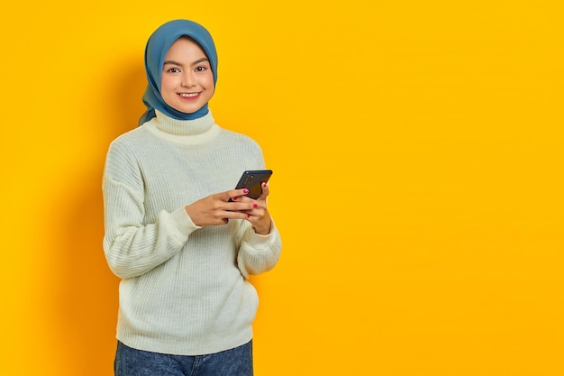 Cheerful young asian woman in white sweater and hijab using on\
mobile phone and looking at camera isolated over yellow background\
people islam religious concept