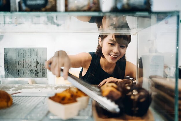 Photo cheerful young asian woman working at a bakery and serving fresh bakery from the display cabinet