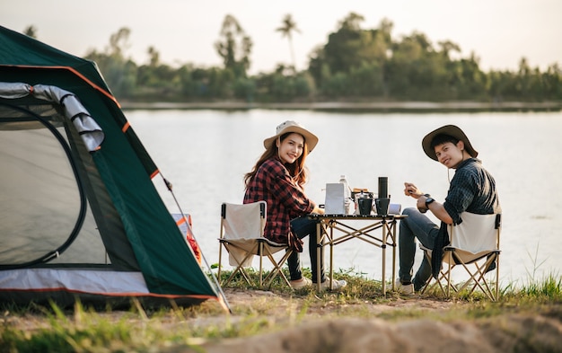 写真 夏休みのキャンプ旅行中にコーヒーセットと淹れたてのコーヒーグラインダーを作る森のテントの前に座っている陽気な若いバックパッカーカップル
