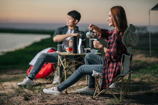 コーヒーセットと森のテントの前に座って、夏休みのキャンプ旅行中に新鮮なコーヒーグラインダーを作る陽気な若いバックパッカーカップル、選択的な焦点