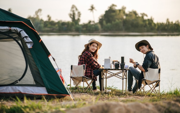 夏休みのキャンプ旅行中にコーヒーセットと淹れたてのコーヒーグラインダーを作る森のテントの前に座っている陽気な若いバックパッカーカップル