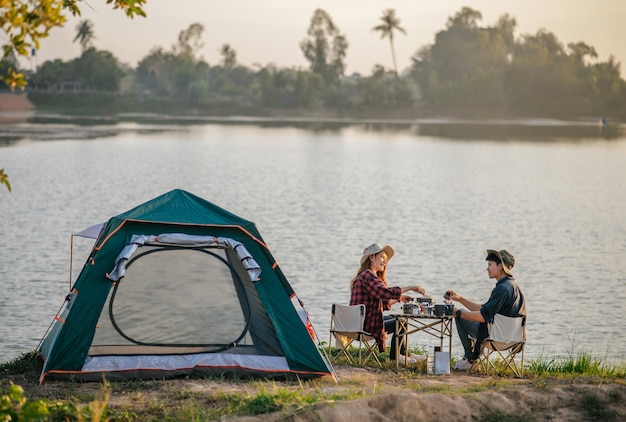 Photo cheerful young backpacker couple wearing trekking hat sitting near lake with coffee and breakfast and making fresh coffee grinder while camping trip on summer vacation