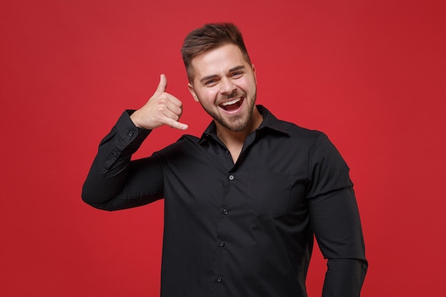 Cheerful young bearded guy 20s in classic black shirt posing isolated on bright red background studio portrait. People lifestyle concept. Mock up copy space. Doing phone gesture says call me back.