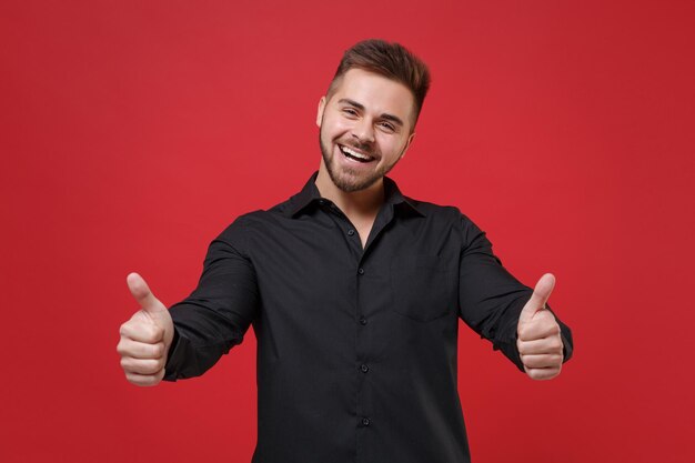 Cheerful young bearded guy 20s in classic black shirt posing isolated on bright red wall background studio portrait. People sincere emotions, lifestyle concept. Mock up copy space. Showing thumbs up.