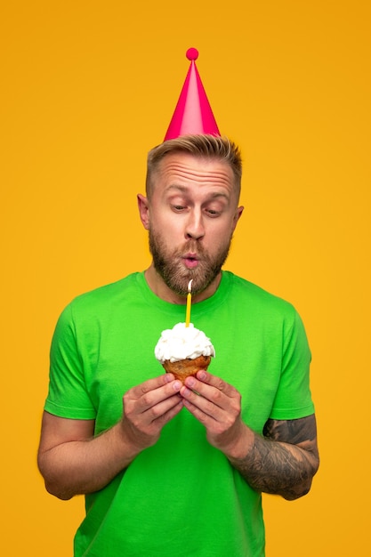 Cheerful young bearded man in bright green shirt and party hat blowing out candle on sweet cupcake with whipped cream during birthday celebration