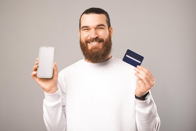 Cheerful young bearded man is holding a phone and card showing screen