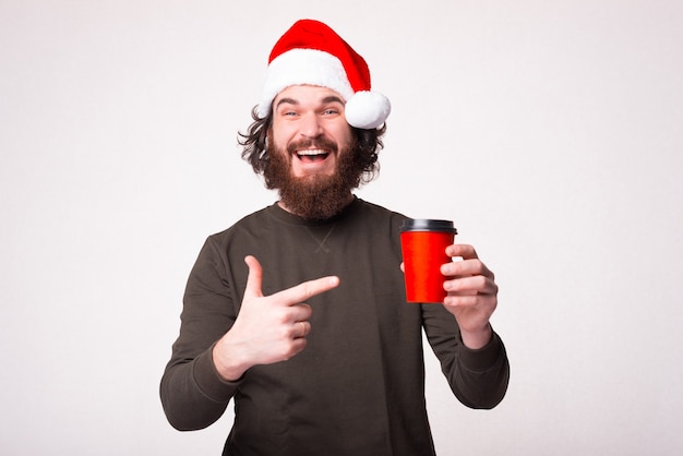 Cheerful young bearded man pointing at red cup of coffee and wearing santa claus hat