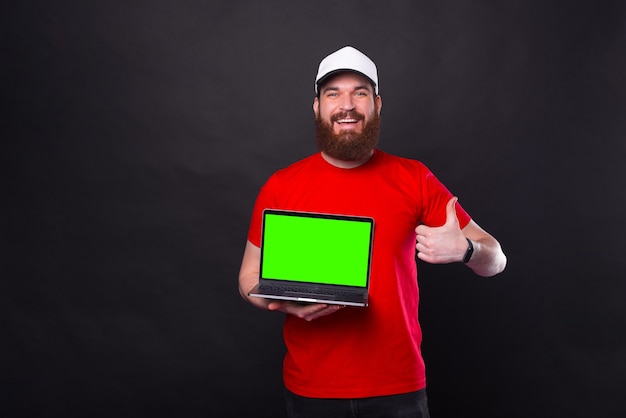 Cheerful young bearded man showing thumb up and green screen on laptop