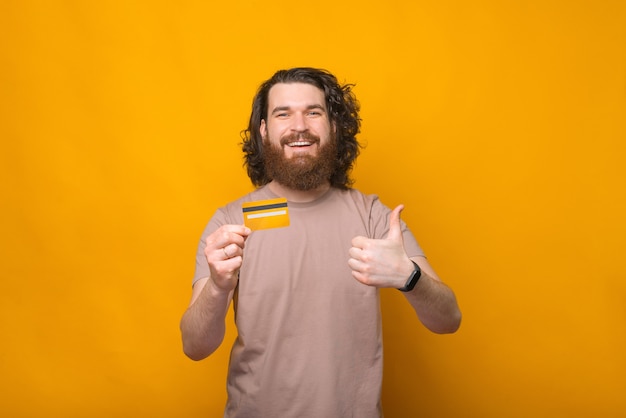 Cheerful young bearded man showing thumb up and his new debit card over yellow