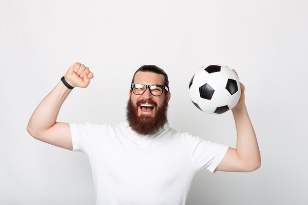 A cheerful young bearded man smiling at the camera is holding a soccer ball near a white wall