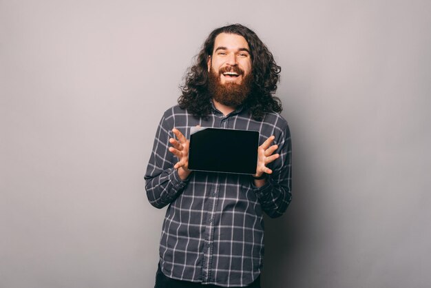 Cheerful young bearded man with long hair showing tablet over grey background