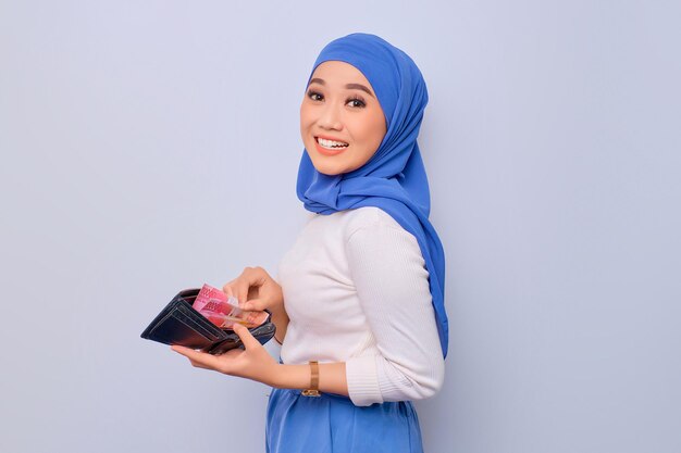 Cheerful young beautiful Asian Muslim woman showing wallet full of money banknotes isolated over white background