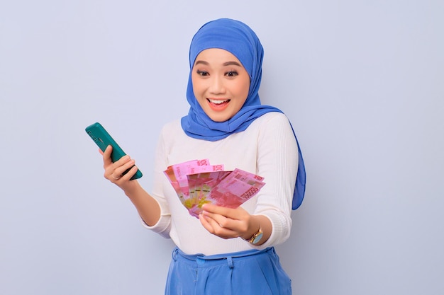 Cheerful young beautiful Asian Muslim woman using mobile phone and holding money banknotes isolated over white background