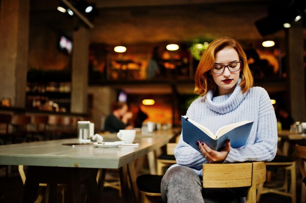 Cheerful young beautiful redhaired woman in glasses sitting at her working place on cafe reading something at her notebook