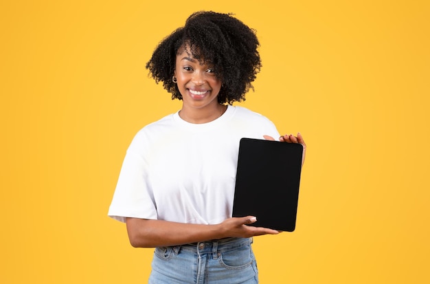 Photo cheerful young black curly lady in white tshirt shows tablet with blank screen isolated on orange