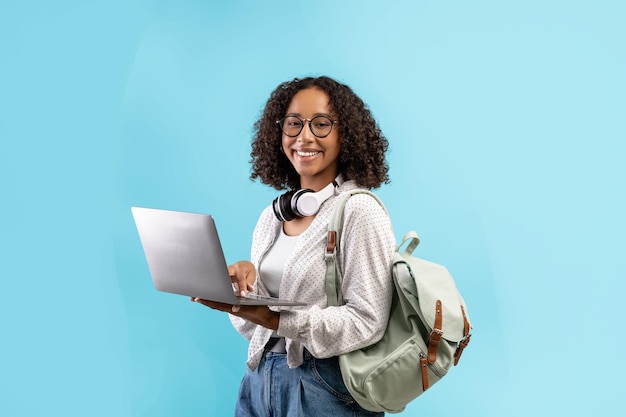 Cheerful young black woman with backpack and headphones holding laptop pc over blue studio