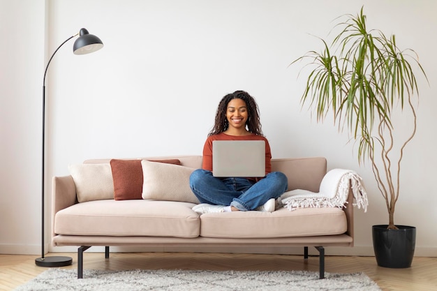 Photo cheerful young black woman working on her laptop while sitting on couch