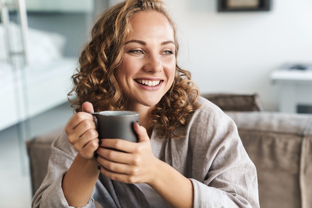 家のソファに座って、目をそらしながらお茶を飲む陽気な若いブロンドの髪の女性