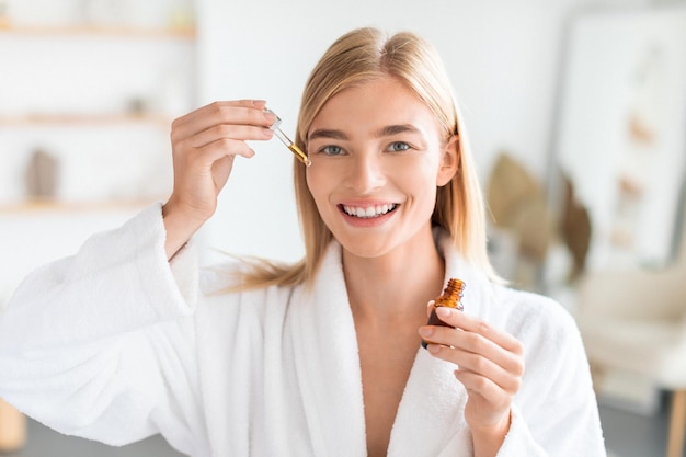 Cheerful young blonde woman applying facial serum moisturizing skin indoor