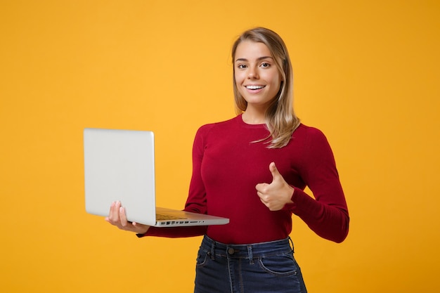 Cheerful young blonde woman girl in casual clothes posing isolated on yellow orange wall background in studio. people lifestyle concept. mock up copy space. hold laptop pc computer, showing thumb up