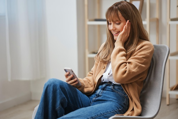 Cheerful young blonde woman in warm sweater get good news on studies by phone touch cheek sitting in armchair at home Pause from work take a break social media in free time concept Copy space