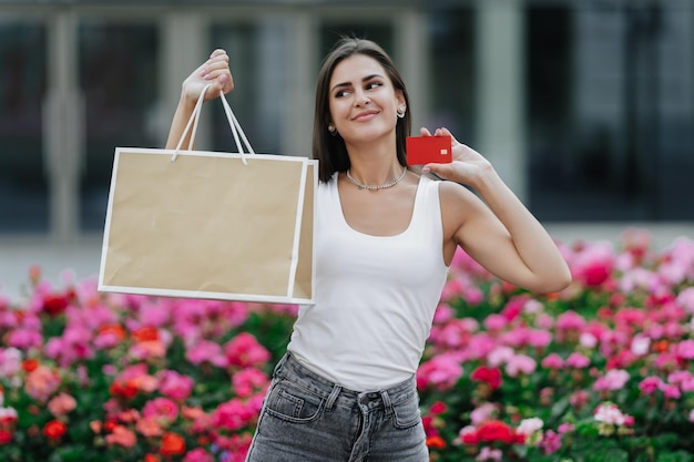 Cheerful young brunette woman in casual holds shopping bag in one hand and credit card by another lo