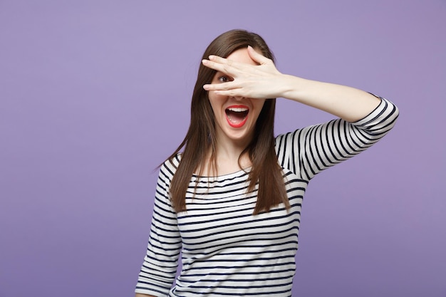 Cheerful young brunette woman in casual striped clothes posing isolated on violet purple background in studio. People lifestyle concept. Mock up copy space. Hiding, peeping, covering eyes with hand.