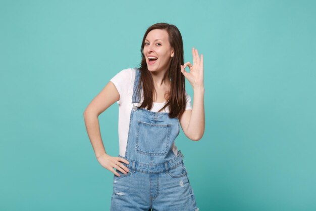 Cheerful young brunette woman girl in casual denim clothes posing isolated on blue turquoise wall background studio portrait. People emotions lifestyle concept. Mock up copy space. Showing OK gesture.