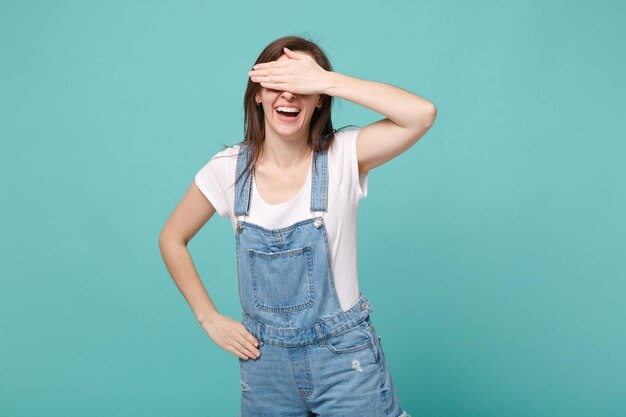 Cheerful young brunette woman girl in casual denim clothes posing isolated on blue turquoise wall background studio portrait. People lifestyle concept. Mock up copy space. Covering eyes with hand.