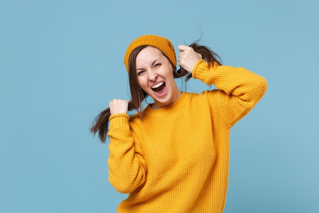 Cheerful young brunette woman girl in yellow sweater hat posing isolated on blue background studio portrait. People sincere emotions lifestyle concept. Mock up copy space. Holding hair like ponytails.