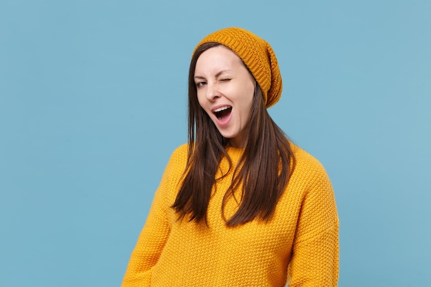 Cheerful young brunette woman girl in yellow sweater and hat posing isolated on blue background studio portrait. People sincere emotions lifestyle concept. Mock up copy space. Looking camera blinking.