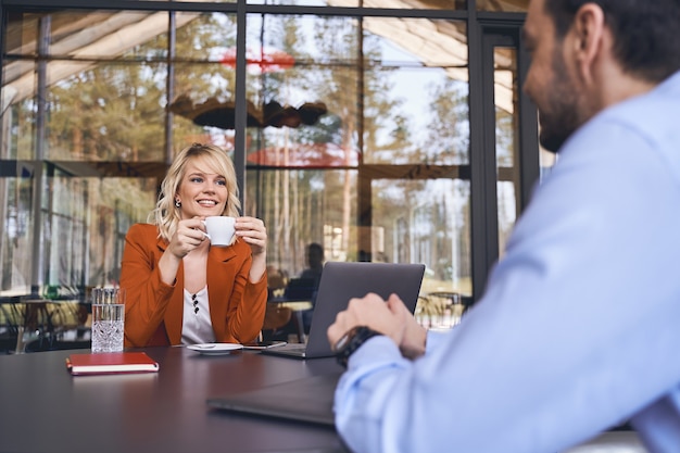 Cheerful young business lady with a cup of tea smiling at her Caucasian male colleague